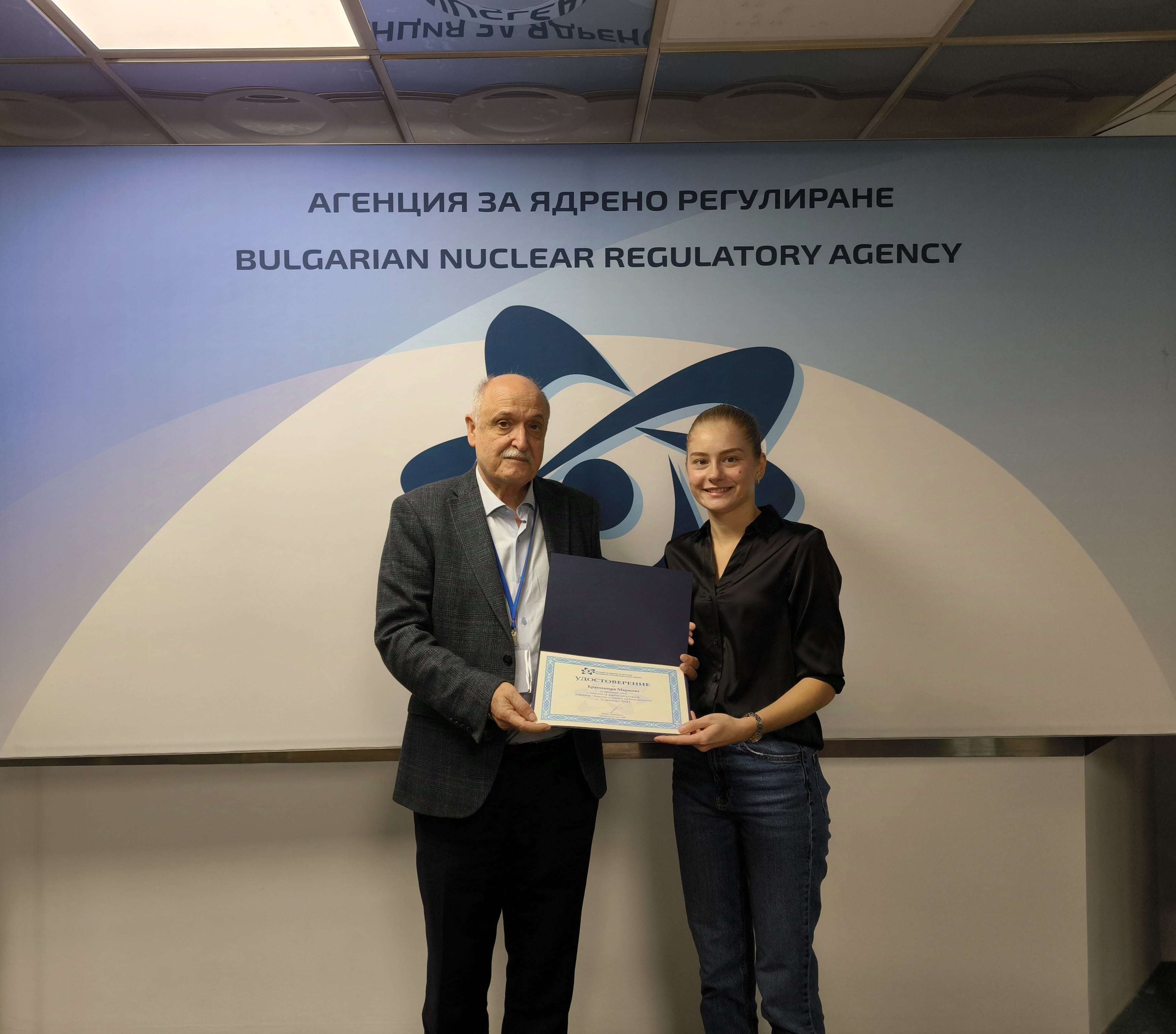 Двама души държащи сертификат пред знак на Агенцията за ядрено регулиране на България.