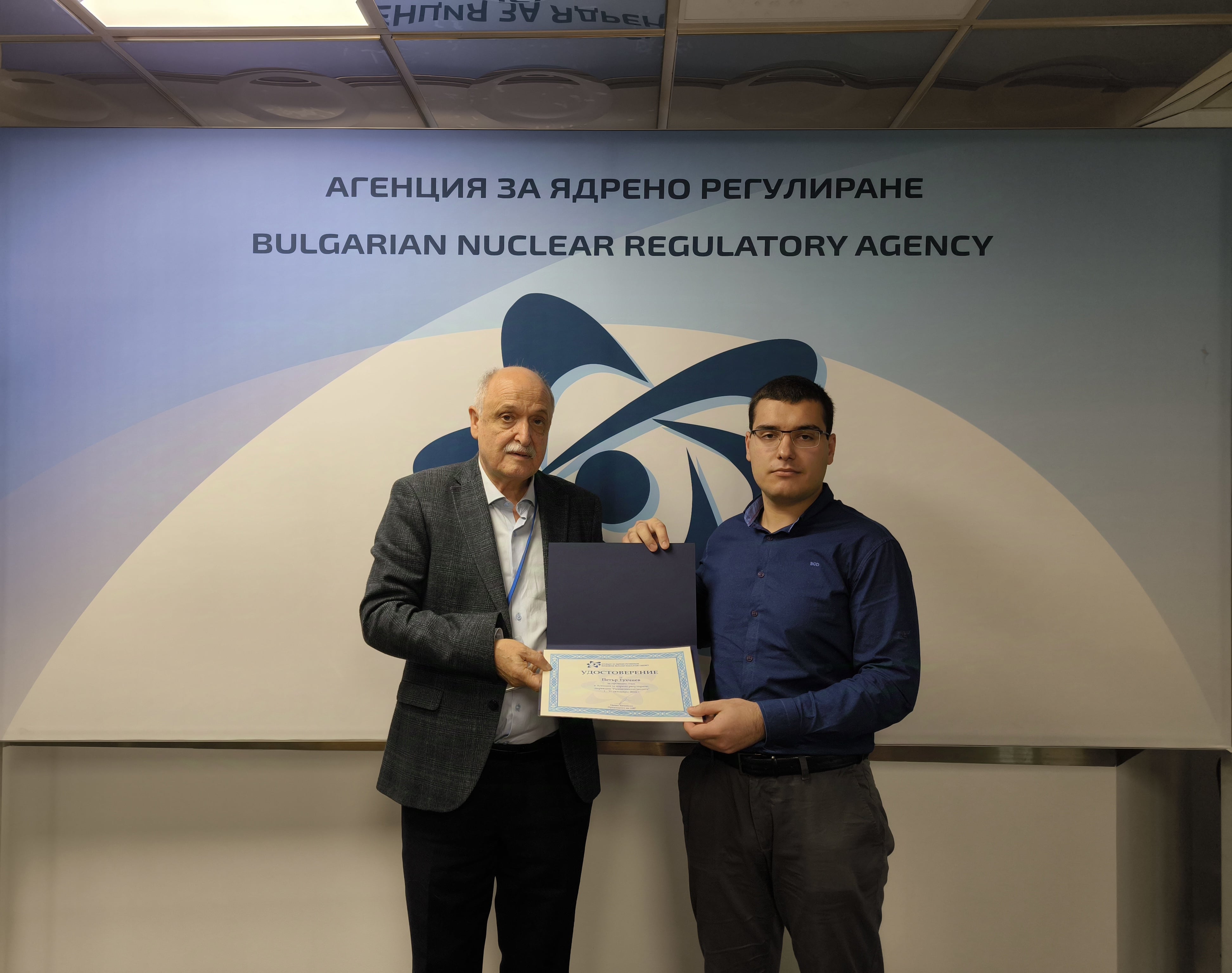 Двама мъже държат сертификат пред табелата на Агенцията за ядрено регулиране на България.