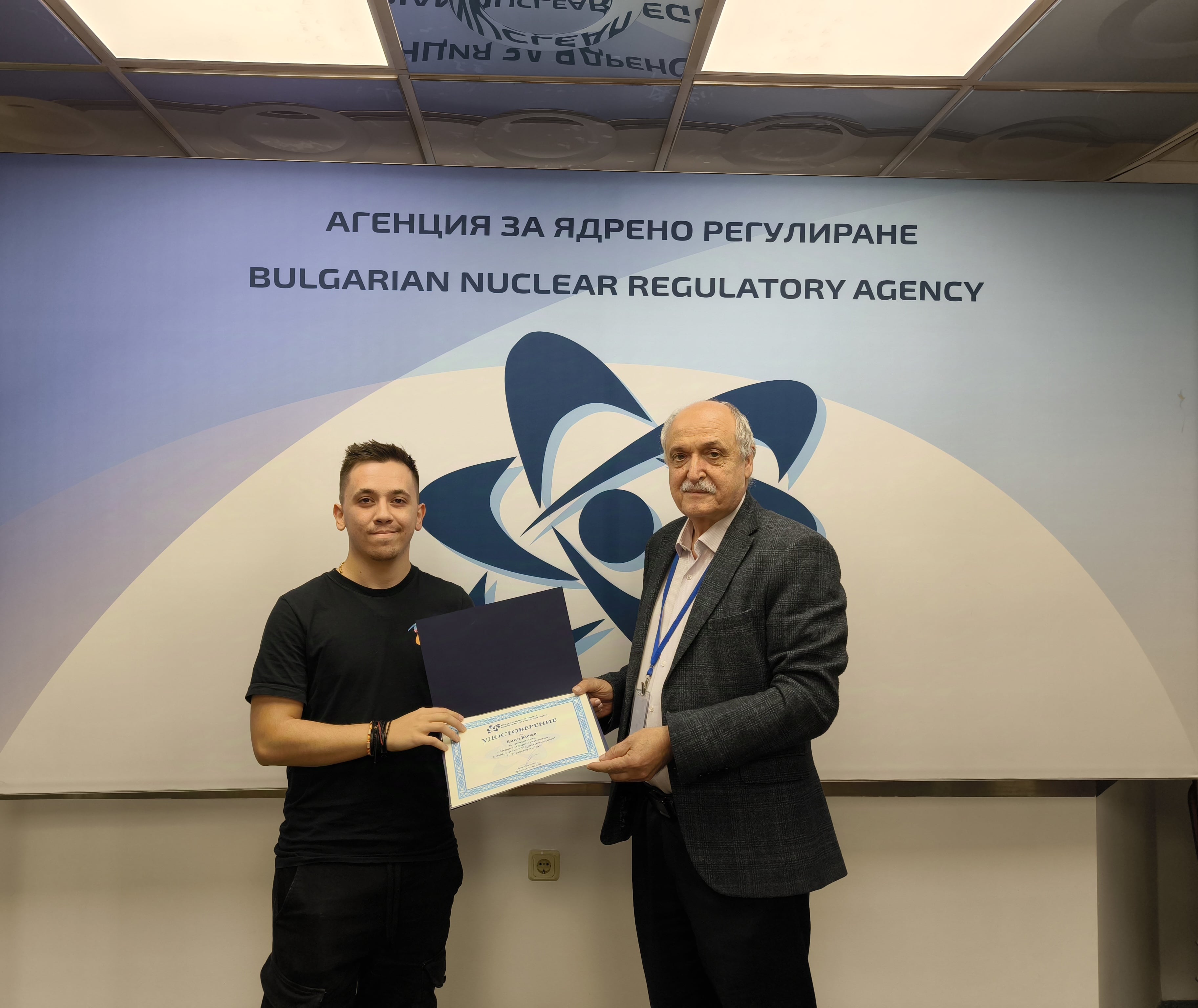Двама мъже стоят, държат сертификат пред табела с надпис "Агенция за ядрено регулиране на България".