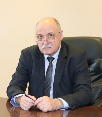 Мъж с очила и мустак, седнал на бюро, облечен в тъмен костюм и вратовръзка, държи химикал.