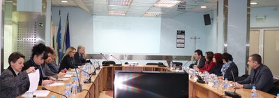 Участници, разположени около П-образна конферентна маса с документи и микрофони в заседателна зала.