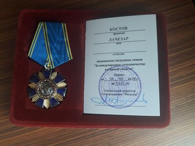 Медал със синя лента в червена кутия, до сертификат на кирилица със син печат.