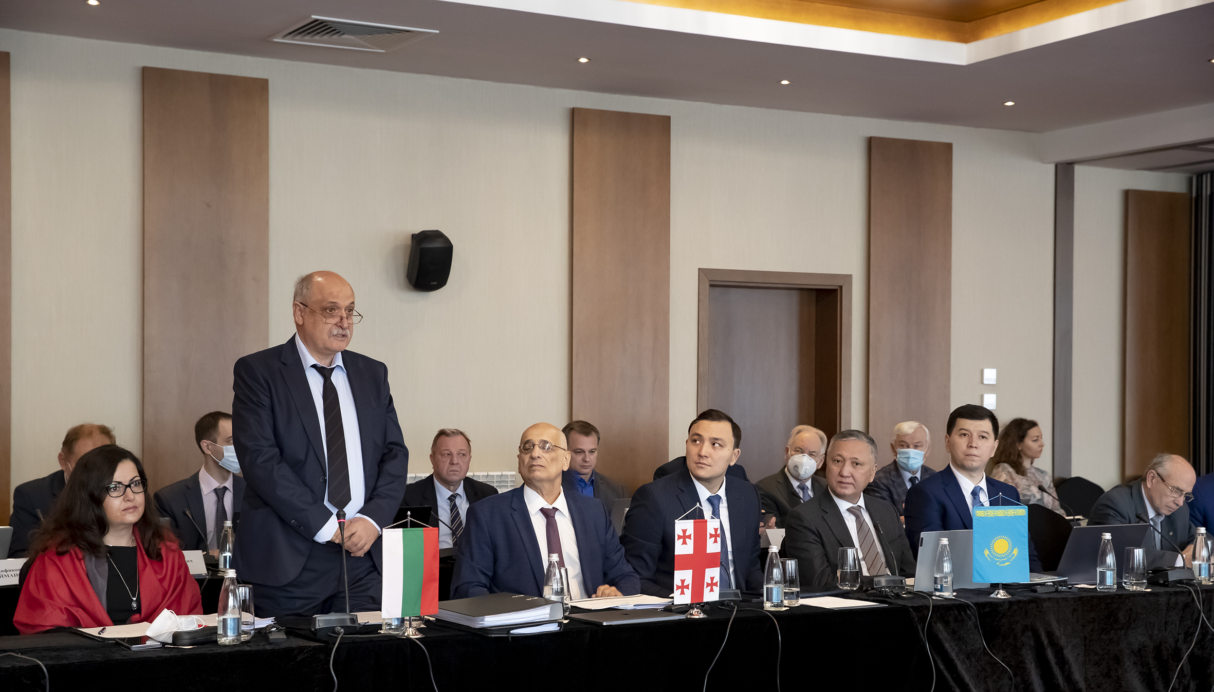 Група лица в официално облекло на конферентна маса с изобразени национални флагове отпред.