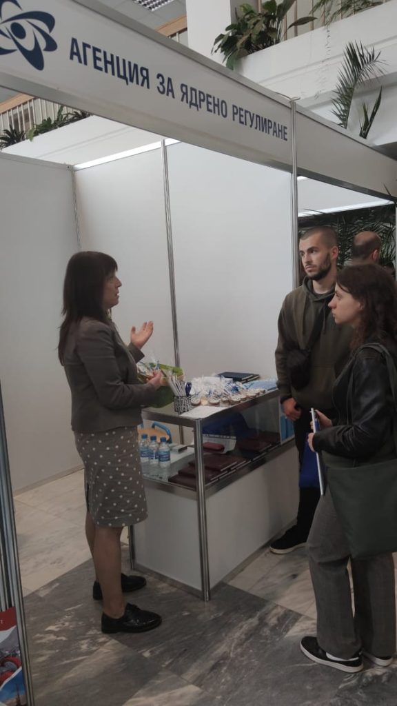 Лице на щанд приветства двама посетители на събитието, организирано от агенцията за ядрено регулиране.