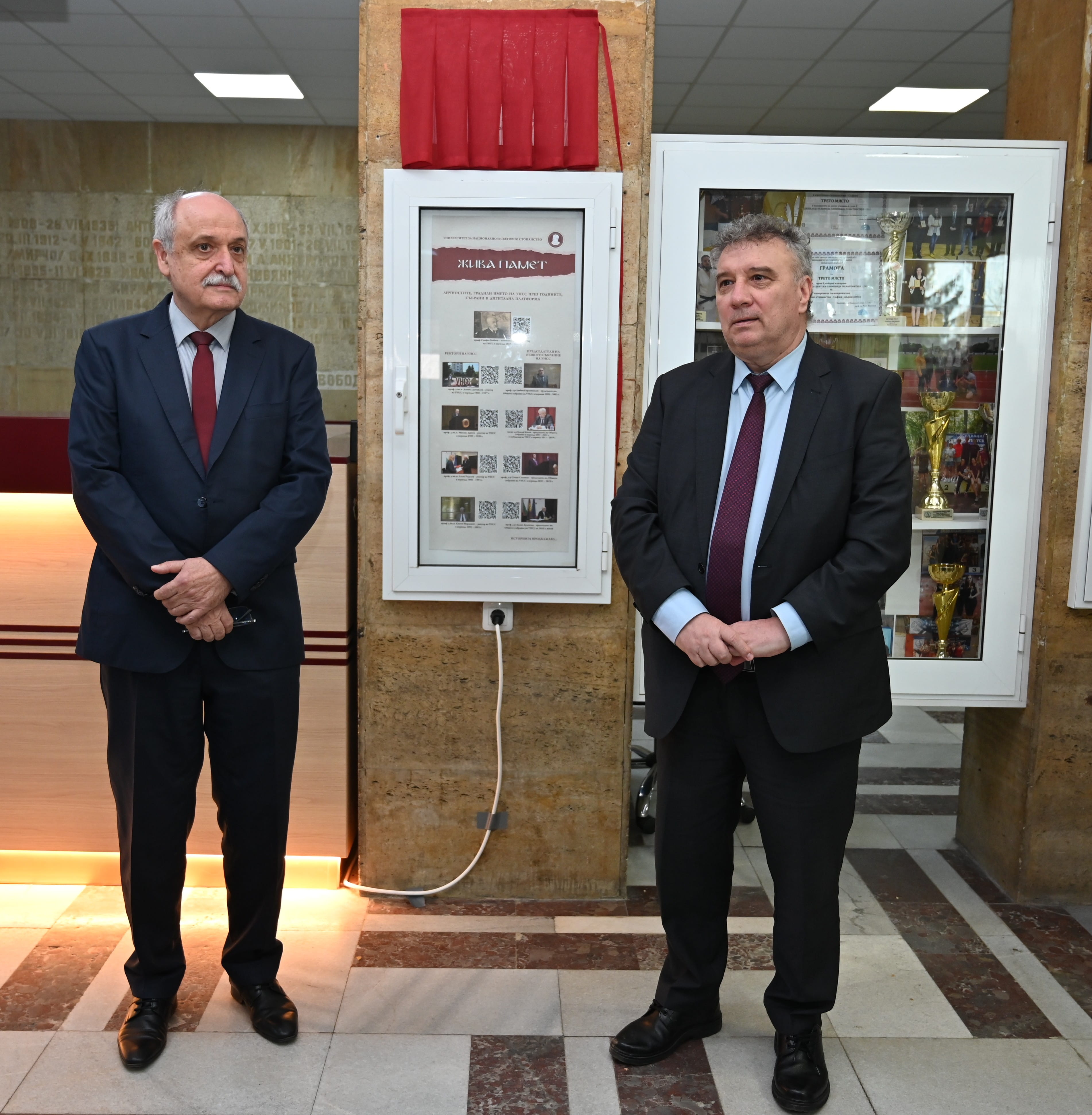 Двама мъже в костюми до витрина с трофеи и плакет с червен плат, на събитие с представители на МААЕ и АЯР.