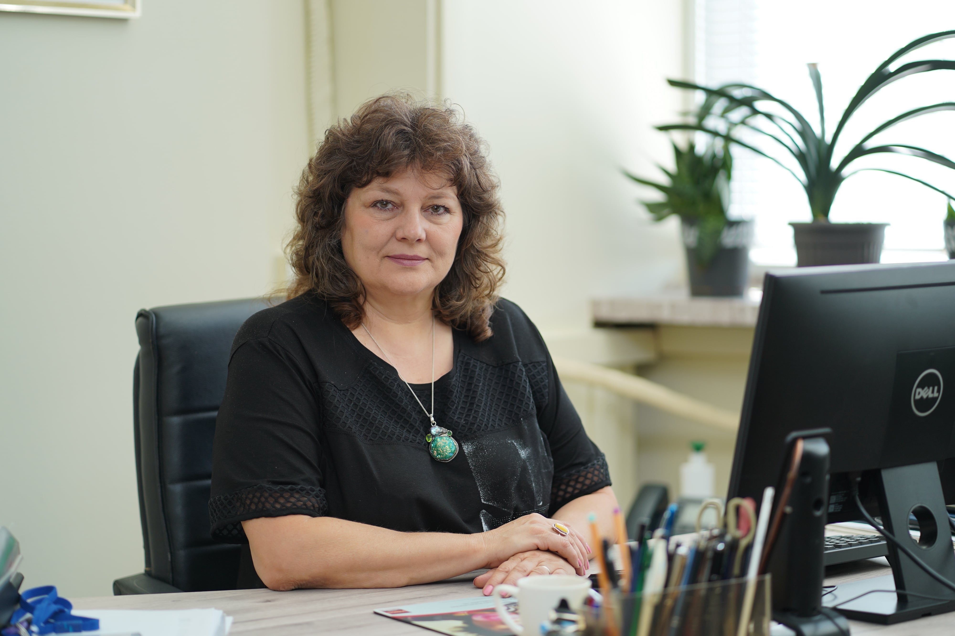 Жена с къдрава коса на бюро в офис. На заден план - растения и компютър.