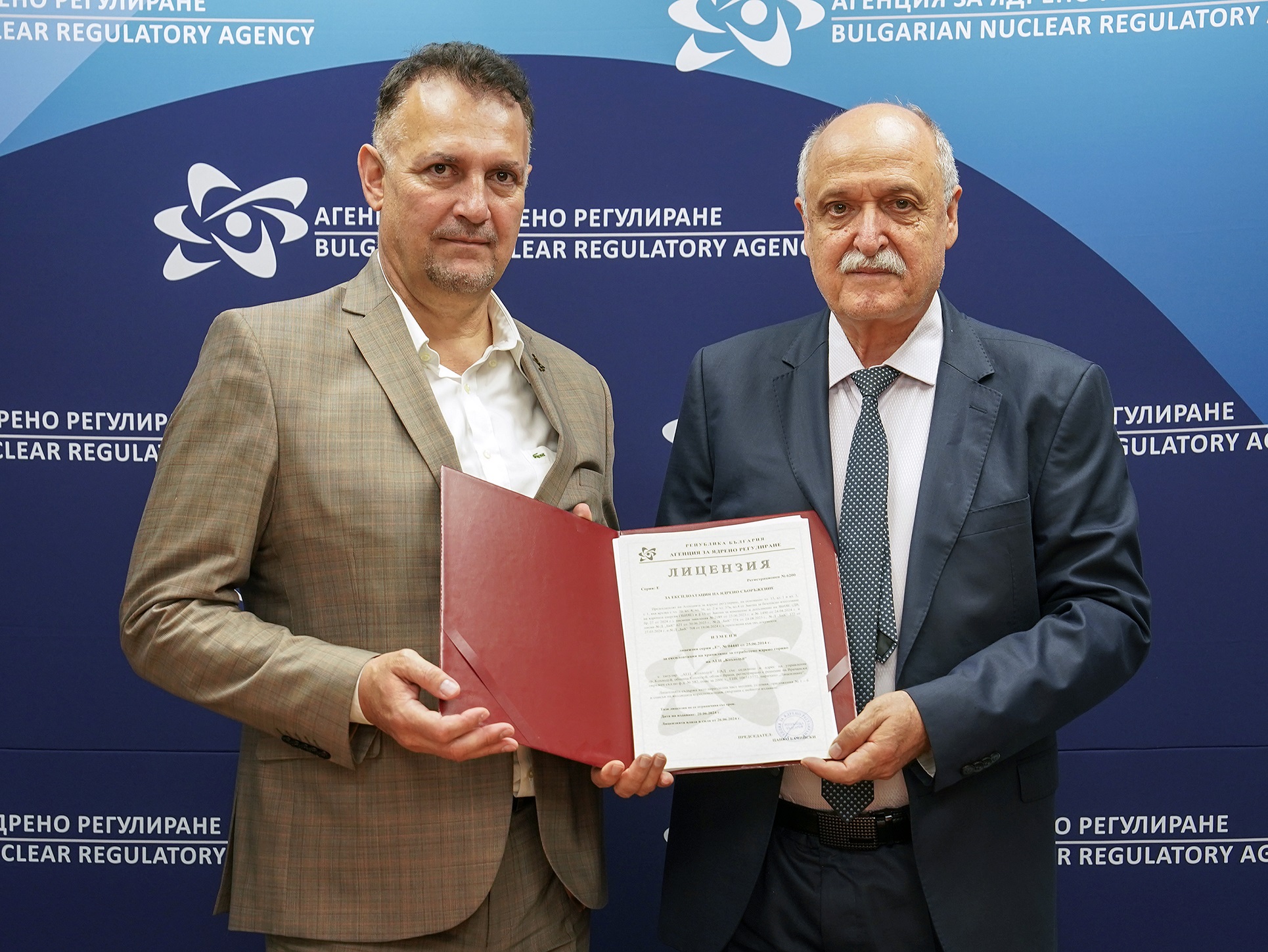 Двама мъже в костюми държат документ пред фон на Българска агенция за ядрено регулиране. Обсъждайте инициативи, свържете се с управлението на отработеното гориво в Хранилище близо до АЕЦ Козлодуй.