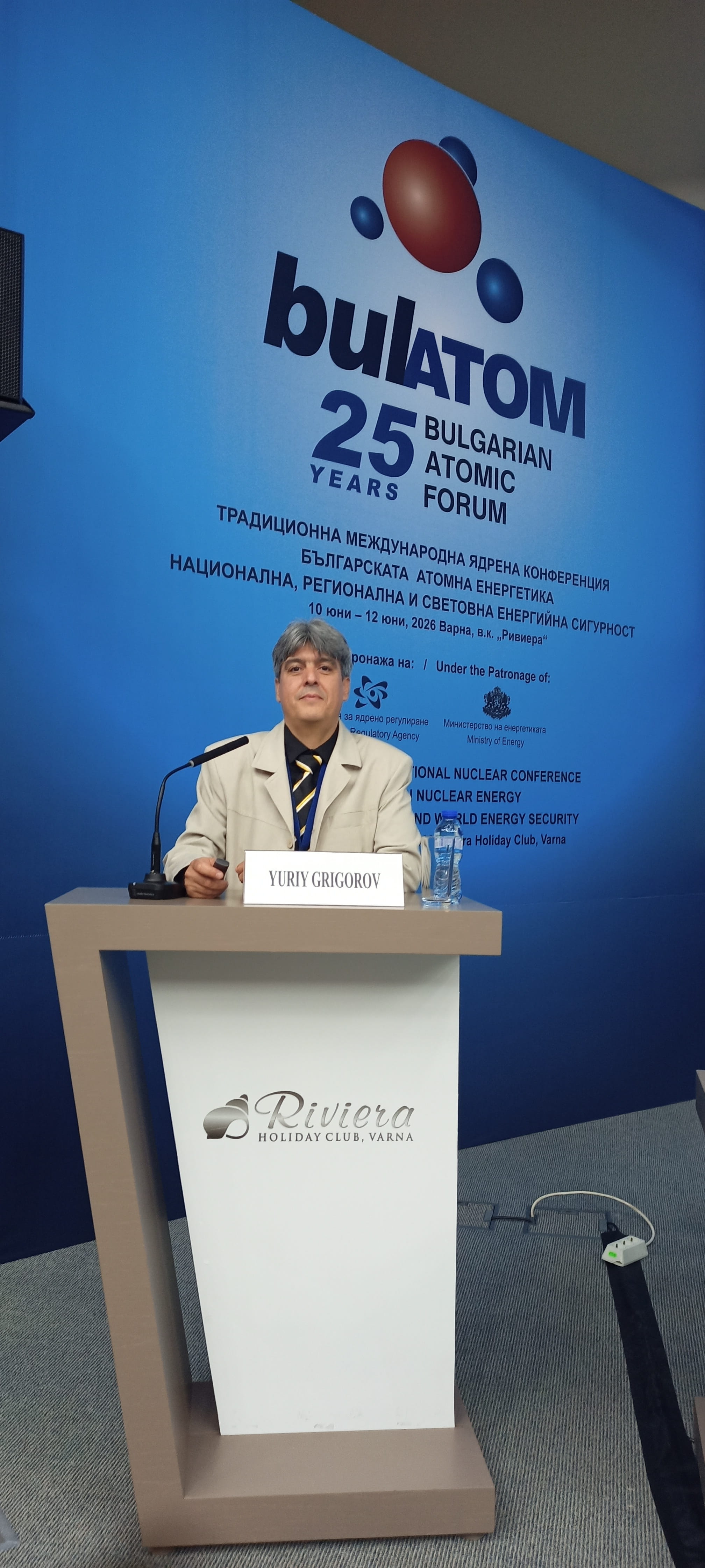 Юрий Григоров, официално облечен, на подиум с надпис името му на 25-та годишнина на БУЛАТОМ ядрената конференция.