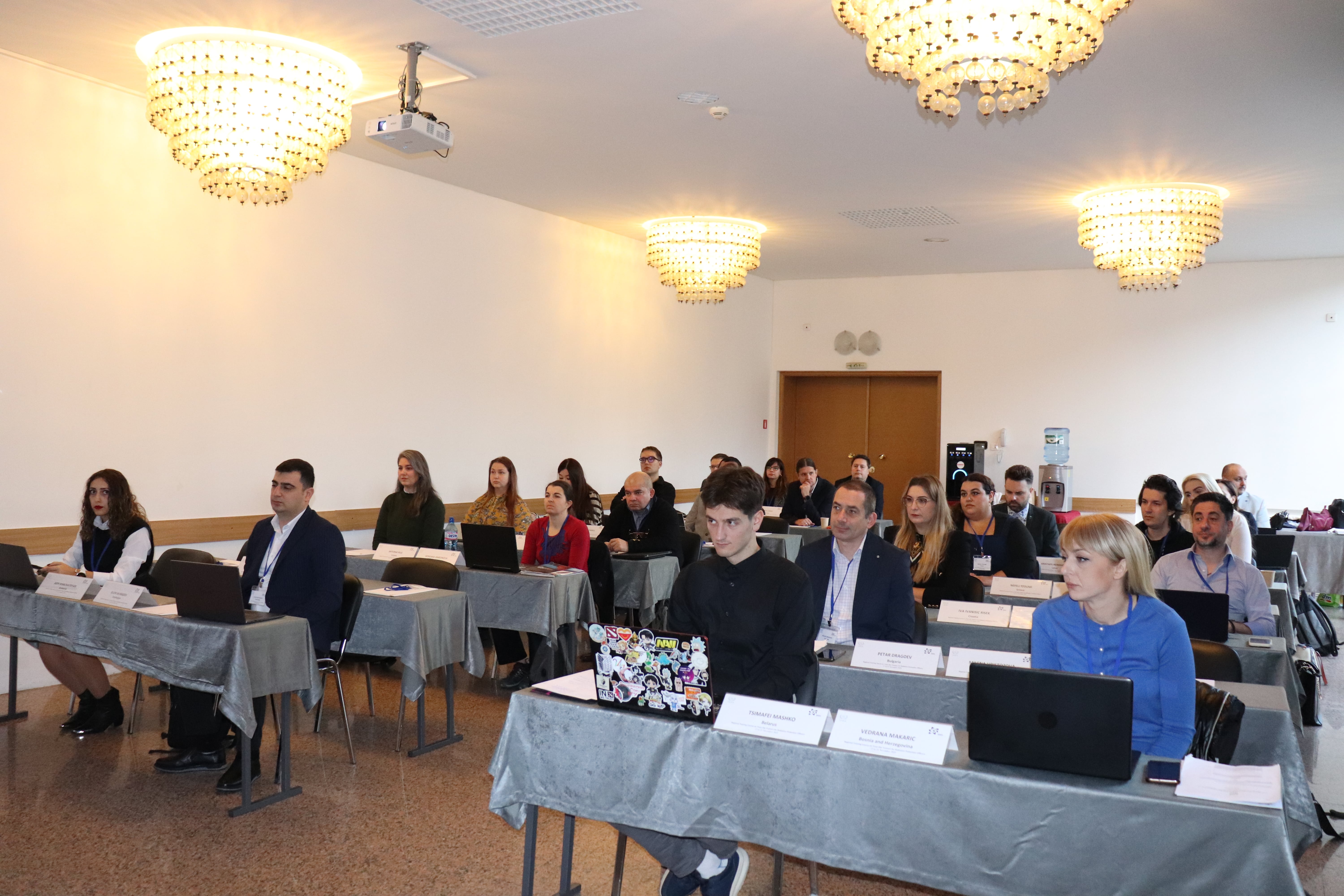 Участници в конферентна зала, прослушват презентация по радиационна защита, работят с лаптопи за повишаване на квалификацията.