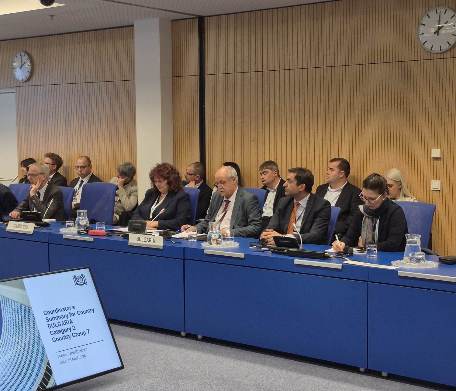 Събрание на АЯР: участници в официално облекло обсъждат ядрена безопасност на маса с табели, блокноти и микрофони.