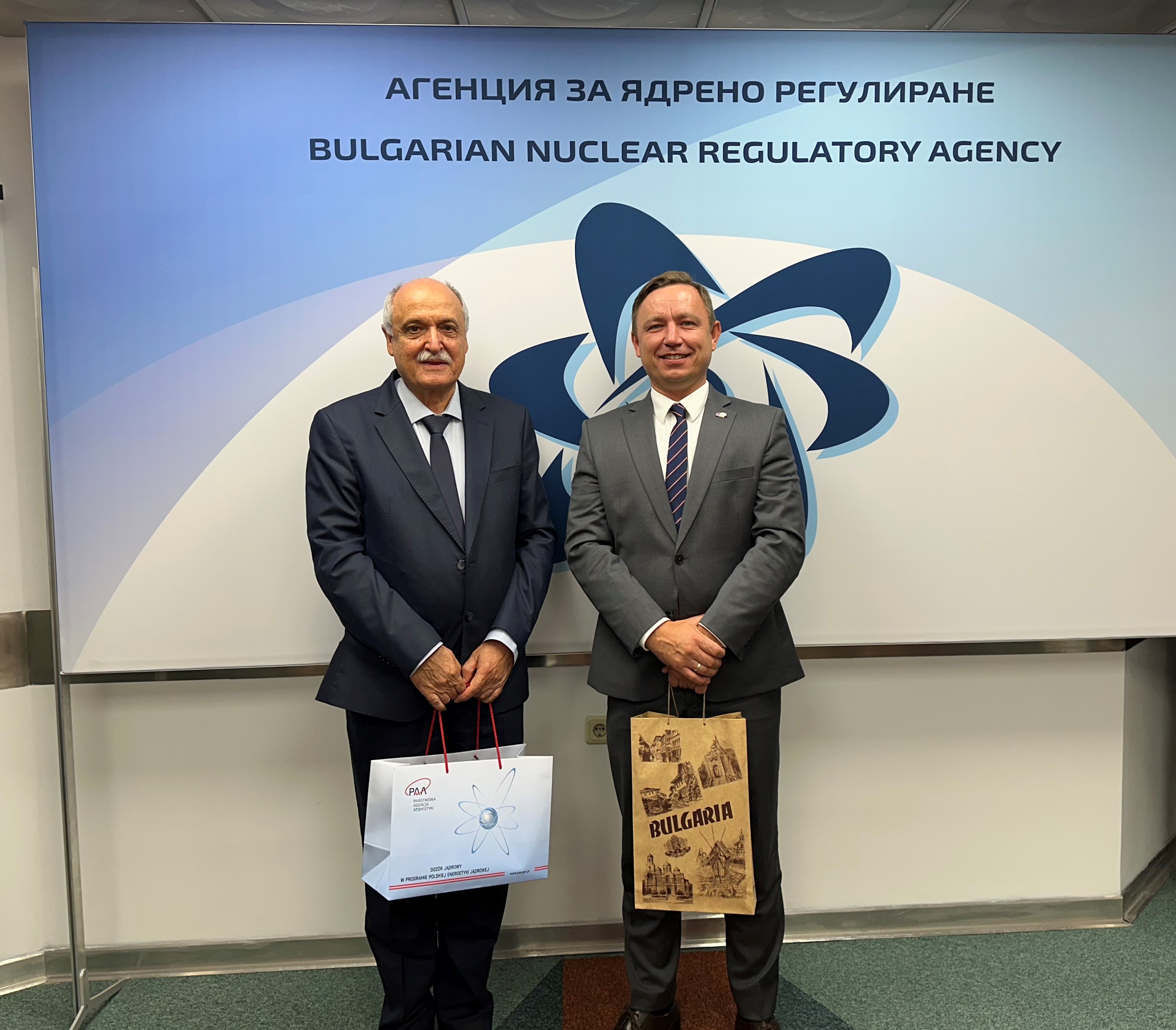 Двама мъже в костюми държат подаръчни торбички пред надпис "Българска агенция за ядрено регулиране".