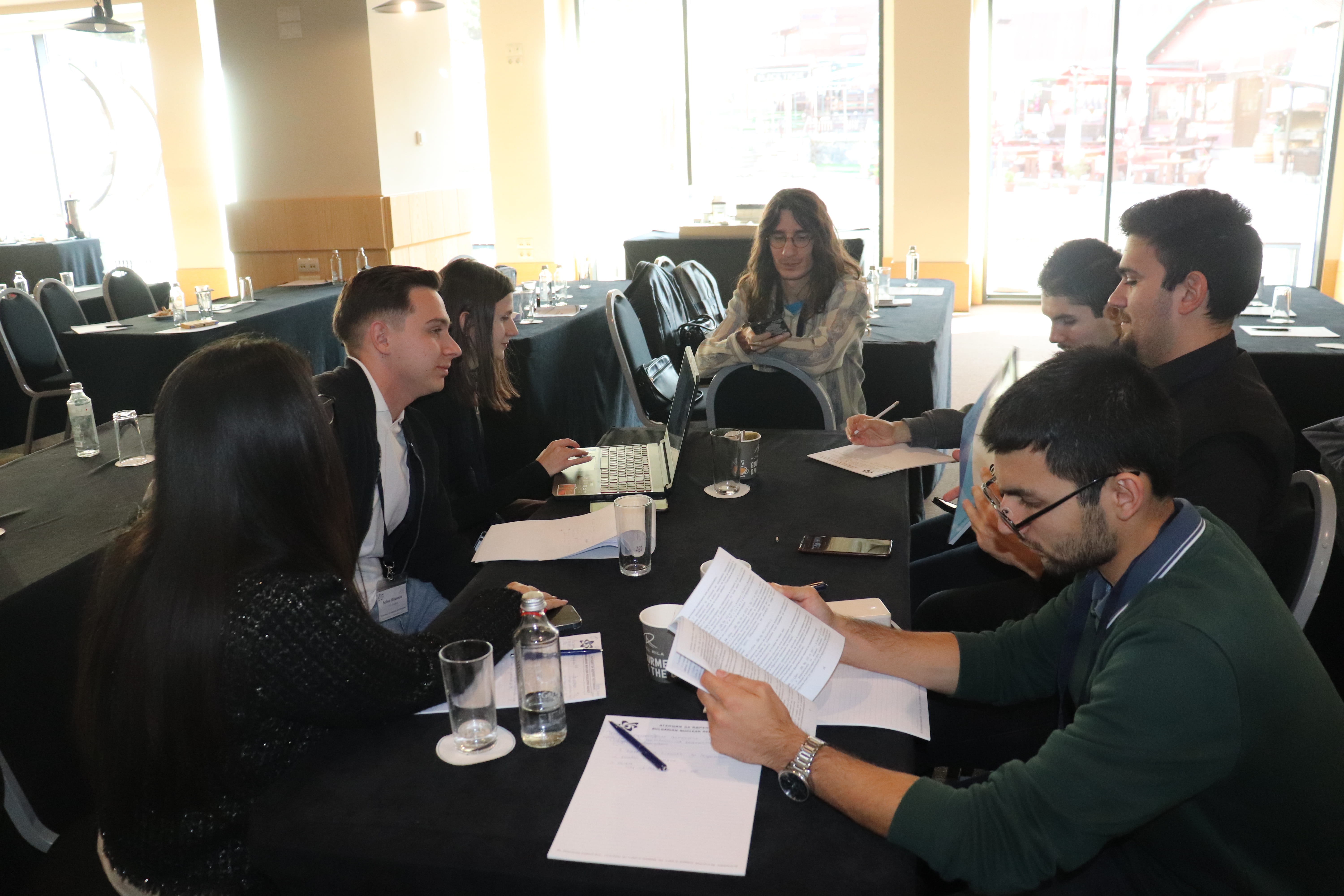 Шестима души заседават в конферентна зала, на маса с документи и лаптопи, обсъждат ядрено регулиране за училищни проекти.