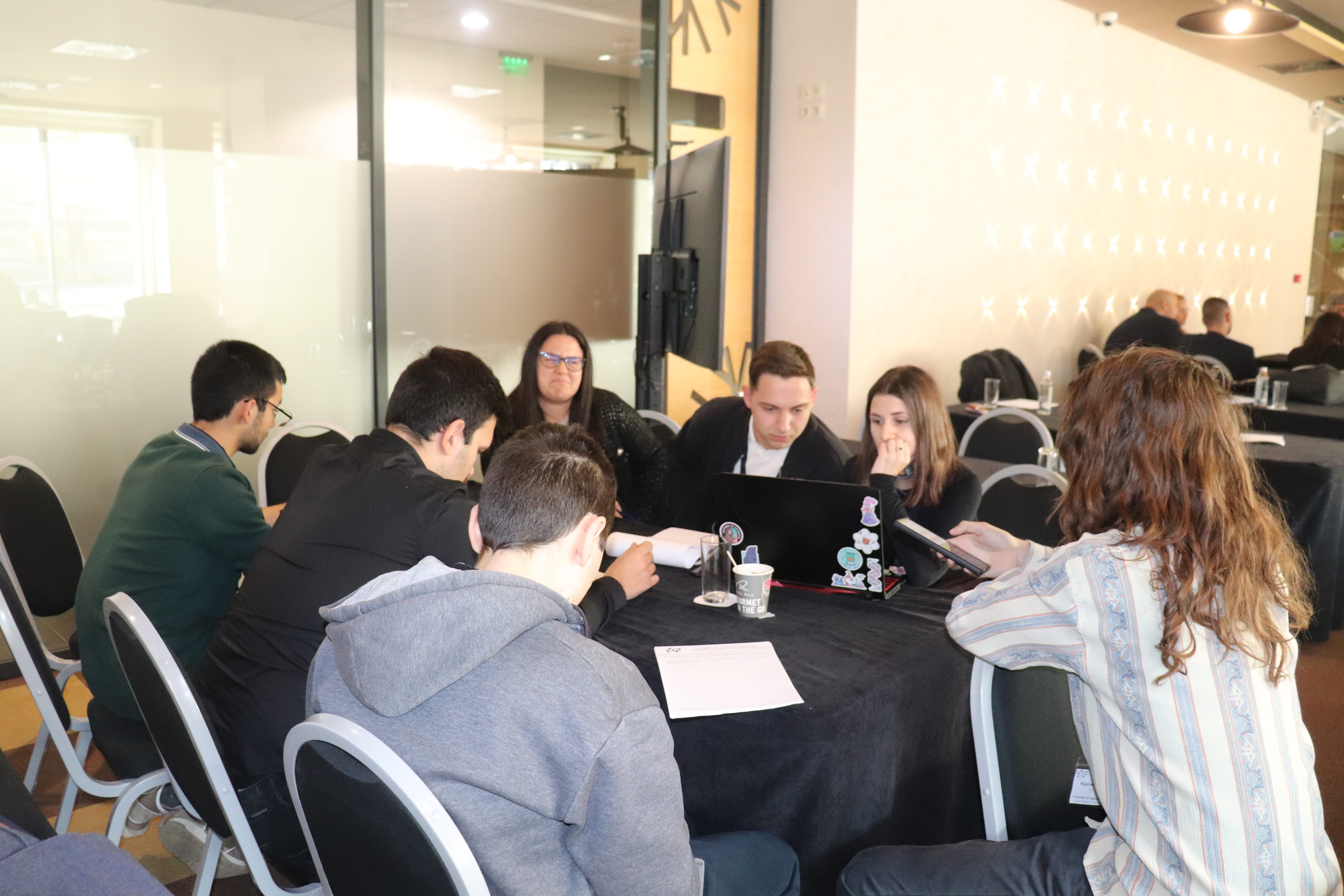 Седем души работят съвместно на маса с лаптопи и документи в модерна, осветена офис среда на училище по ядрено регулиране.