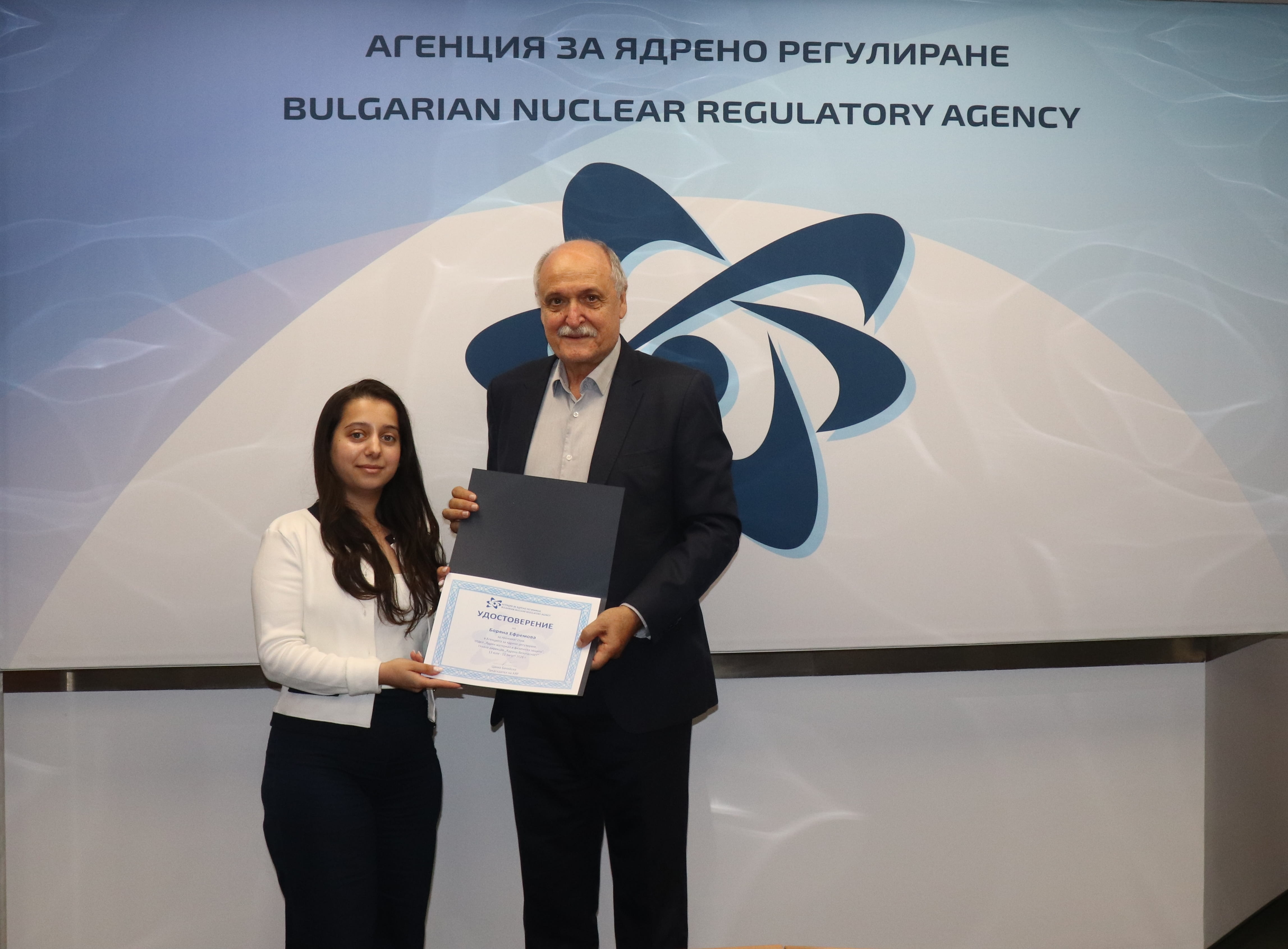 Двама души държат сертификат на фона на АЯР Българска агенция за ядрено регулиране по повод успешно завършения платен летен стаж на студентите-участници.