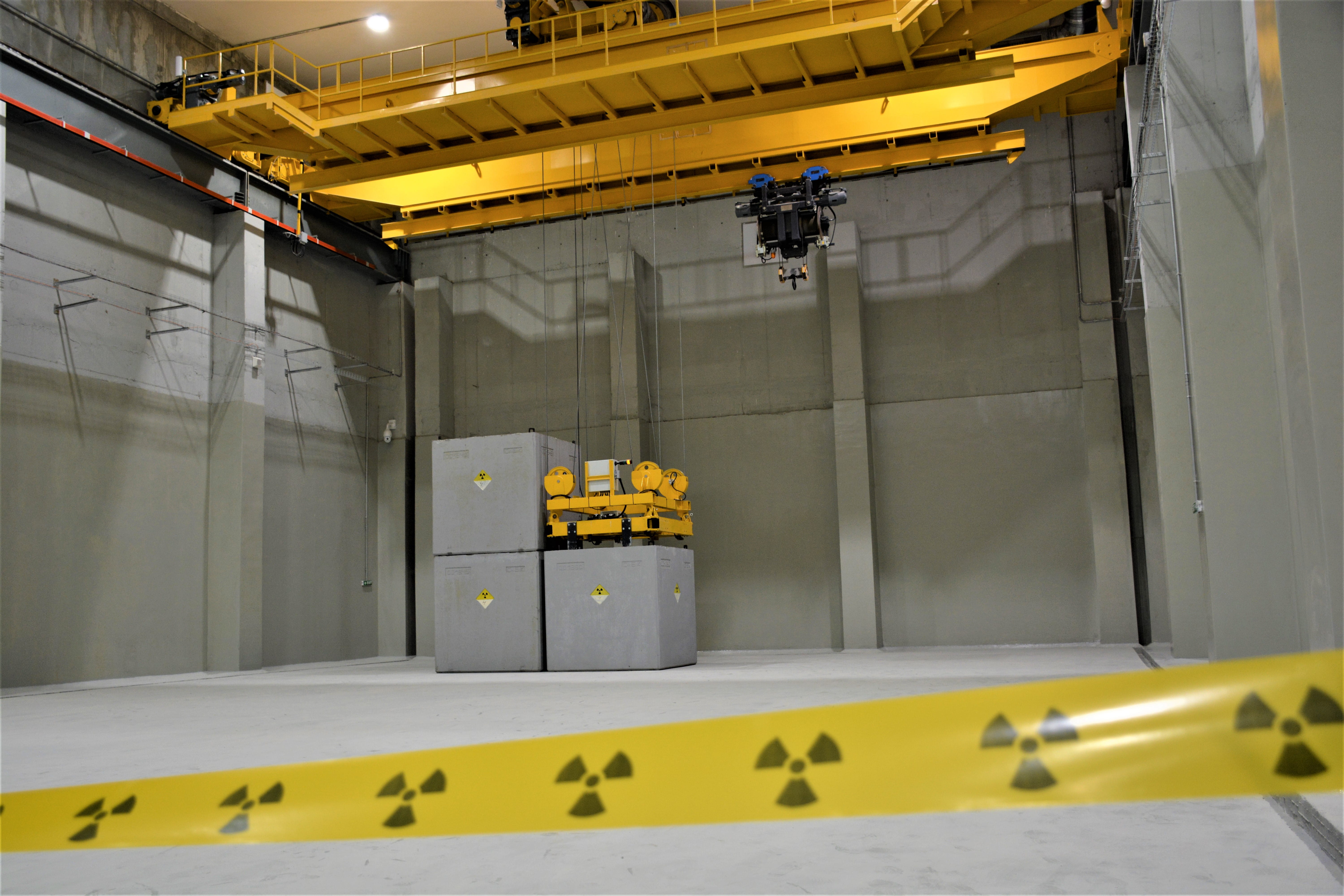 В сигурно бетонно помещение са разположени големи сиви контейнери; отпред има жълта лента с радиационно предупреждение.