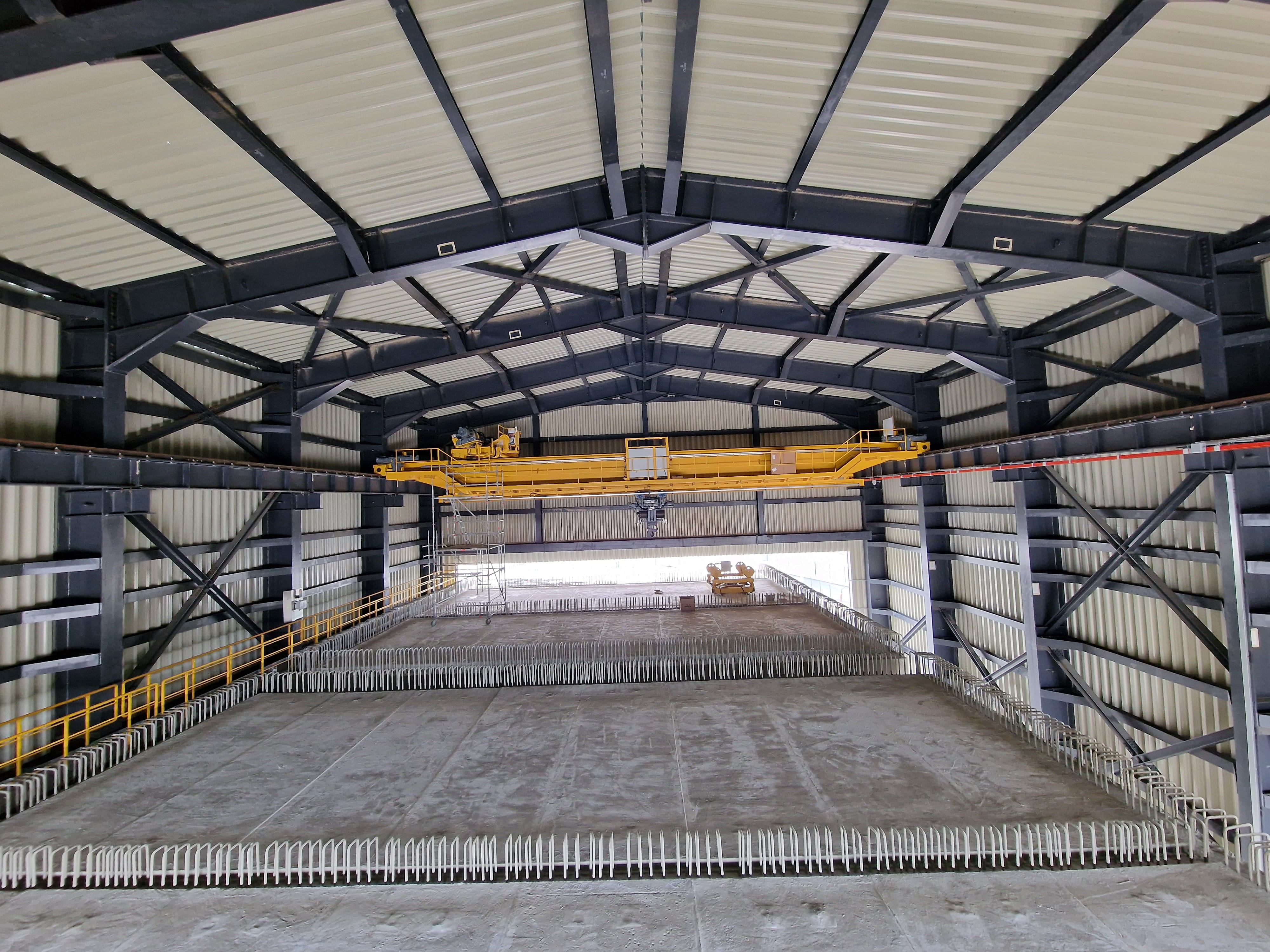 Интериор на голям индустриален склад с метални греди, жълт мостов кран и празен бетонен под.