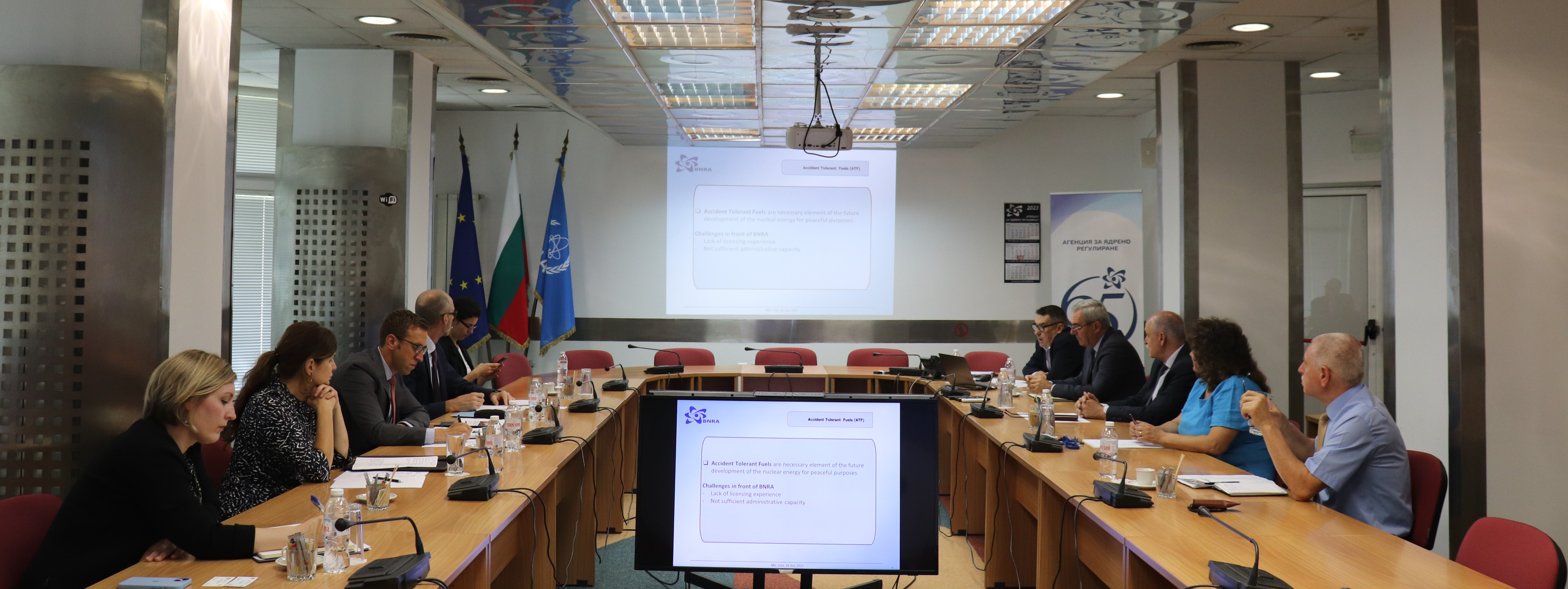 Участници, заседаващи около конферентна маса с презентация и знамена в зала за срещи.
