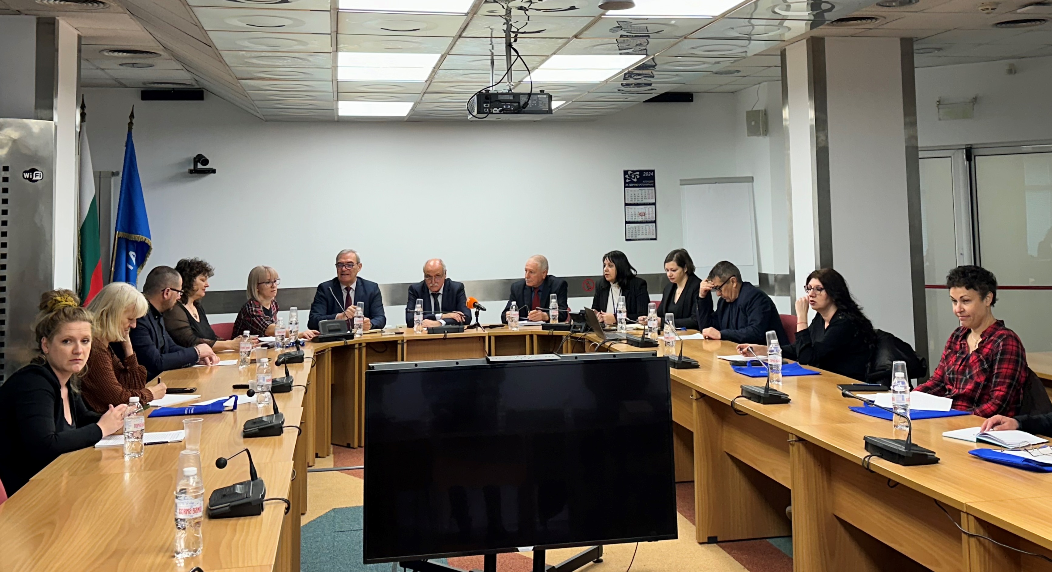 Група лица заседават около голяма конферентна маса с документи, участници във формално съвещание.